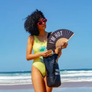 A person in a yellow swimming costume and red sunglasses stands on the beach, holding the "I'm hot!" black fan—an effortlessly curated look inspired by concept store style, with the ocean and blue sky as a backdrop. Gate21.ee