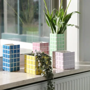 A yellow Planter Tile (14 x 14 x 14 cm)—an example of Estonian design—sits on a windowsill holding a plant. Sunlight streams through the window, casting geometric shadows beside it. Gate21.ee