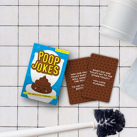 A pack of Brain Training while you Poo - Cards rests on white bathroom tiles beside two cards, a toilet brush, and a glimpse of toilet paper—perfect for gift shop shelves or anyone seeking unique brain teasers for the loo. Gate21.ee