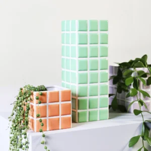 The Vase Tile - Mint (10.5 x 10.5 x 28 cm) features a stylish grid pattern and holds lush green plants, making it an eye-catching choice for modern gift shop displays against light-coloured backgrounds. Gate21.ee