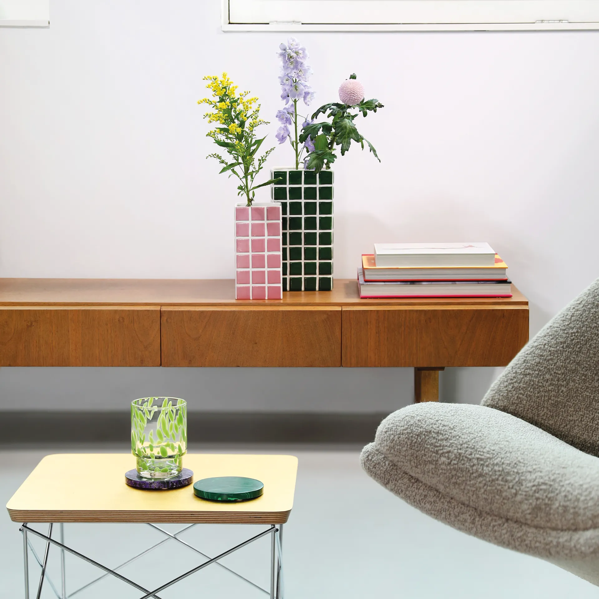 A modern living room features a curated wooden sideboard with stacked books and the Vase Tile – Pink (10.5 x 10.5 x 21 cm) holding flowers. In front, a yellow table displays two green glasses, next to a grey textured chair. Gate21.ee