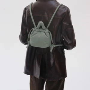 A person in a dark brown leather jacket and trousers, hair tied back, faces away with the Micro Ona Soft Bag - Sage—an ideal accessory for finding unique ideas at a concept shop. The background is plain and light-coloured. Gate21.ee