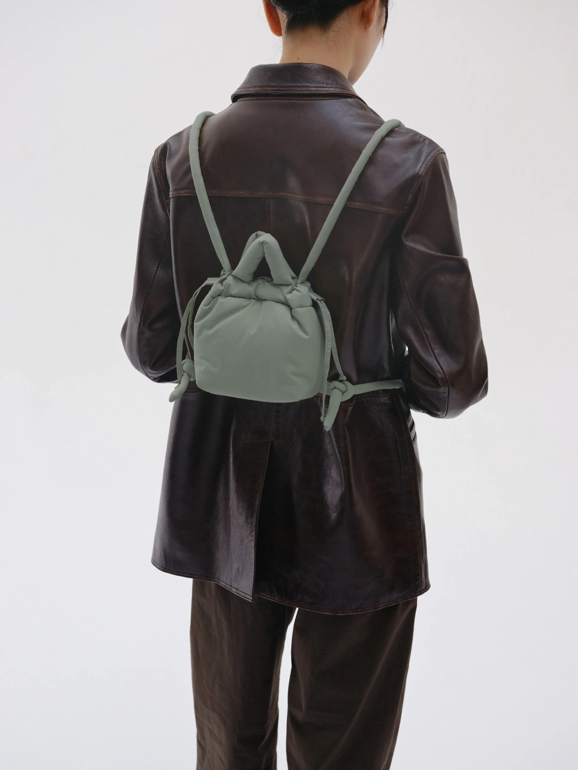 A person in a dark brown leather jacket and trousers, hair tied back, faces away with the Micro Ona Soft Bag - Sage—an ideal accessory for finding unique ideas at a concept shop. The background is plain and light-coloured. Gate21.ee