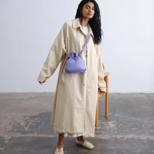 A person stands indoors on a concrete floor, effortlessly styled in a long beige trench coat, cream shoes, and the Micro Ona Soft Bag in Lilac—a nod to refined Estonian minimalist design. Gate21.ee