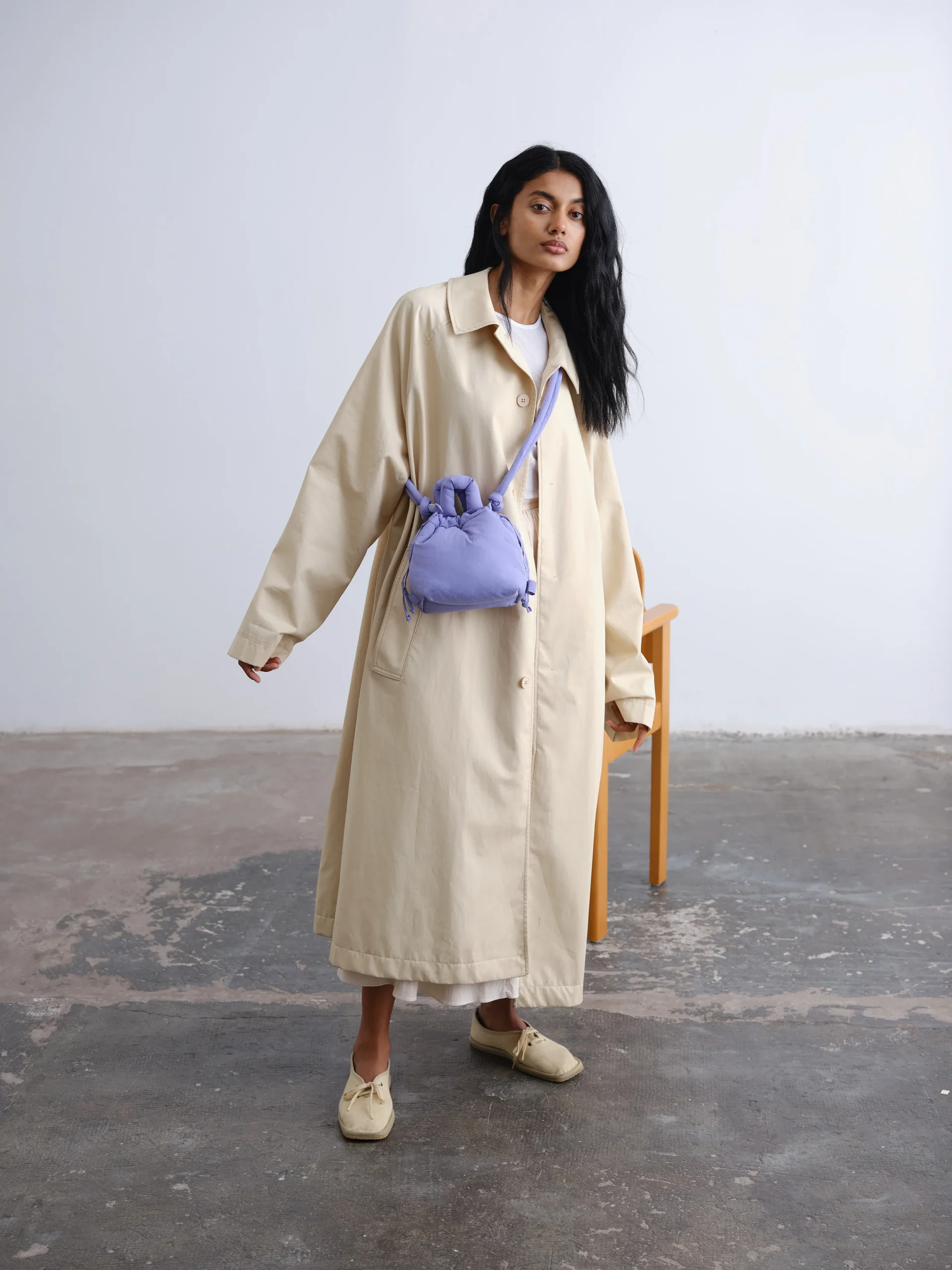 A person stands indoors on a concrete floor, effortlessly styled in a long beige trench coat, cream shoes, and the Micro Ona Soft Bag in Lilac—a nod to refined Estonian minimalist design. Gate21.ee