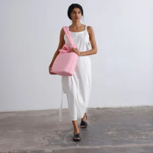 A person wearing a sleeveless white dress and black sandals walks forward, holding the Ona Soft Bag in pale pink against a plain white wall and concrete floor. Gate21.ee