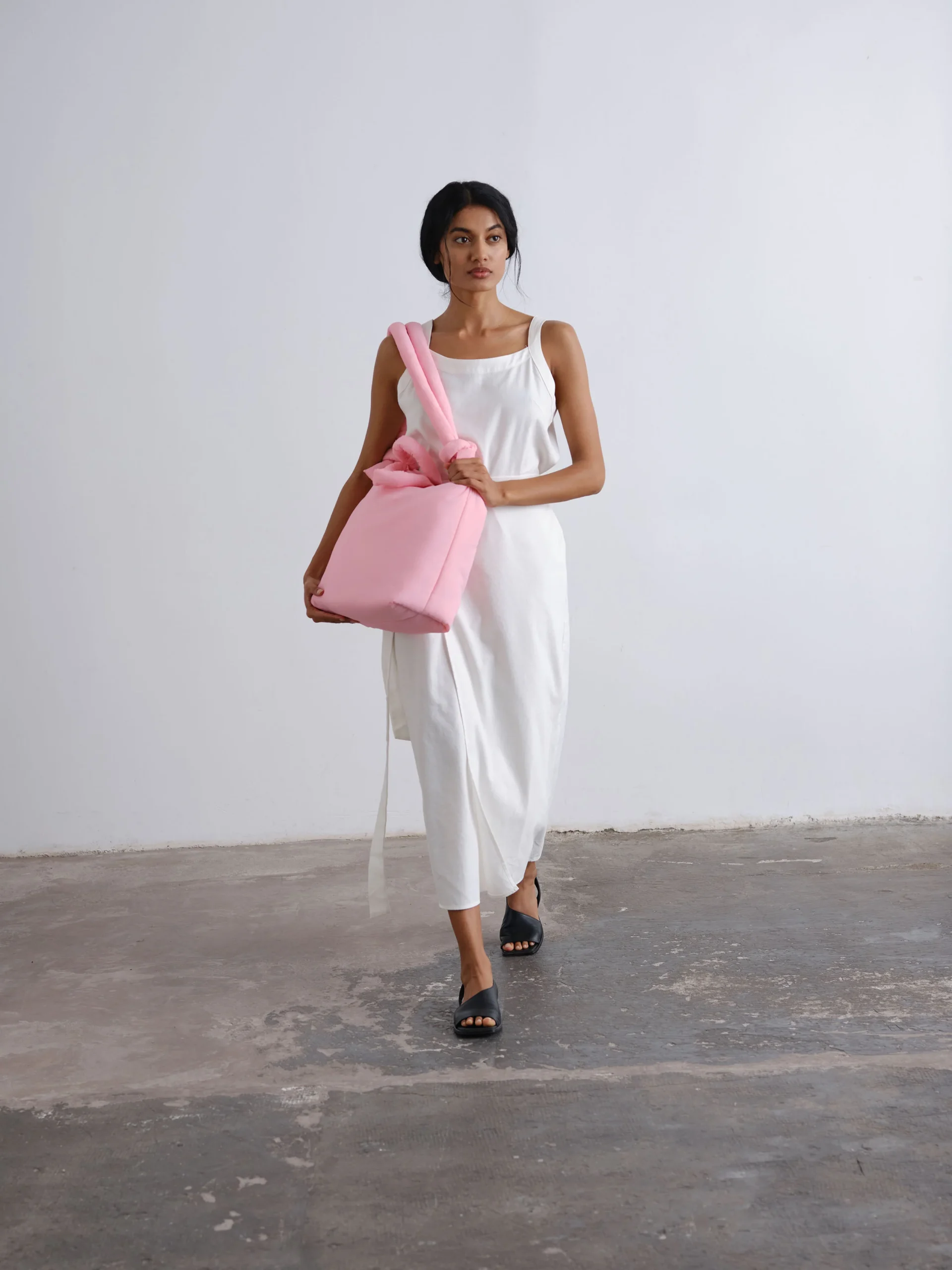 A person wearing a sleeveless white dress and black sandals walks forward, holding the Ona Soft Bag in pale pink against a plain white wall and concrete floor. Gate21.ee