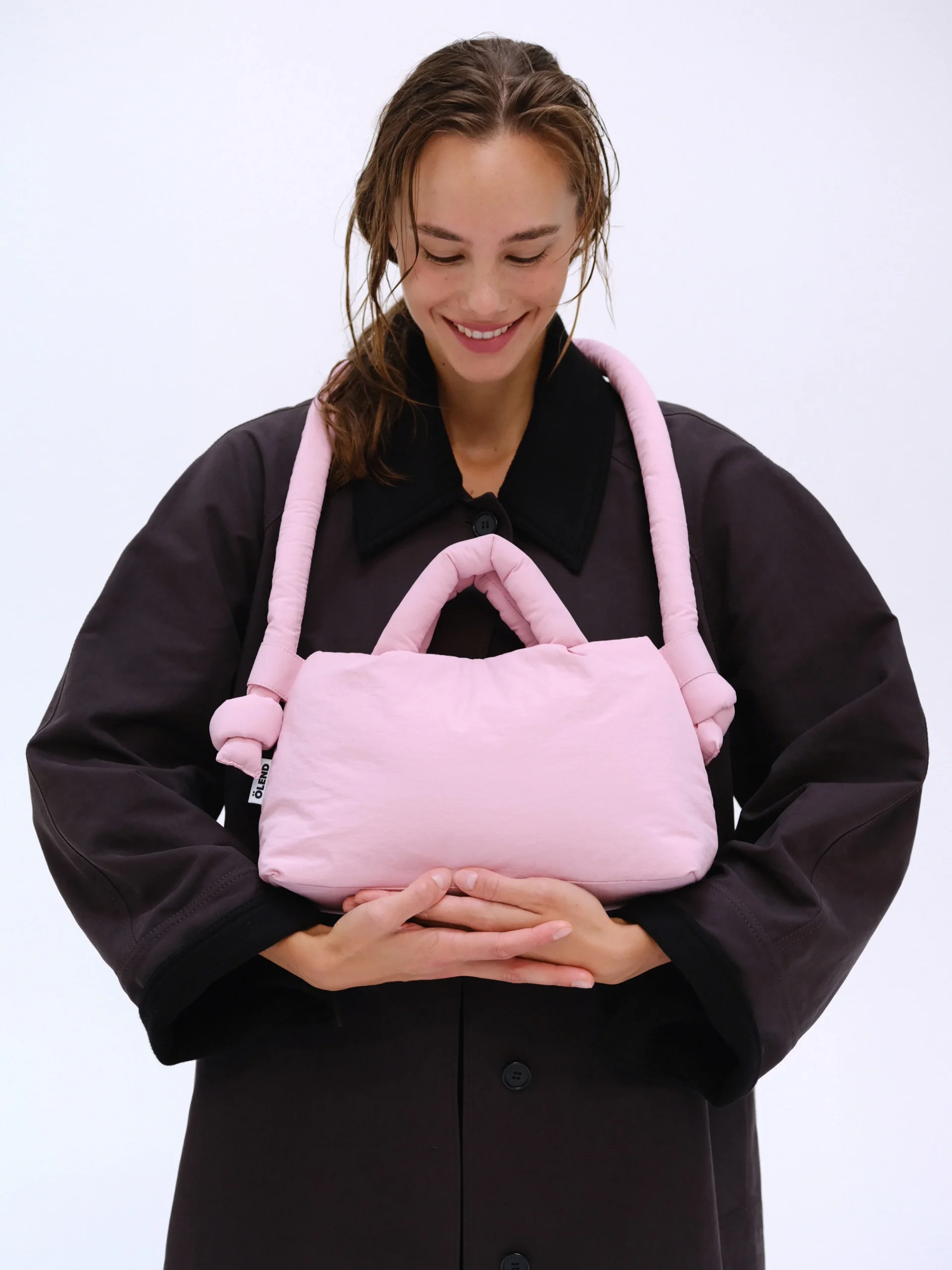 A person in a dark oversized coat holds and gazes at the Mini Ona Soft Bag in pale pink, featuring two looped handles, against a plain white background—a curated concept-store-inspired accessory. Gate21.ee