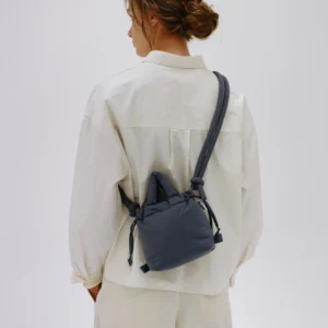 A person with brown hair in a low messy bun, wearing a loose off-white shirt and trousers, stands facing away from the camera. They carry the Micro Ona Soft Bag - Coal, perfect for stylish Tallinn shopping or exploring Estonian design. Gate21.ee