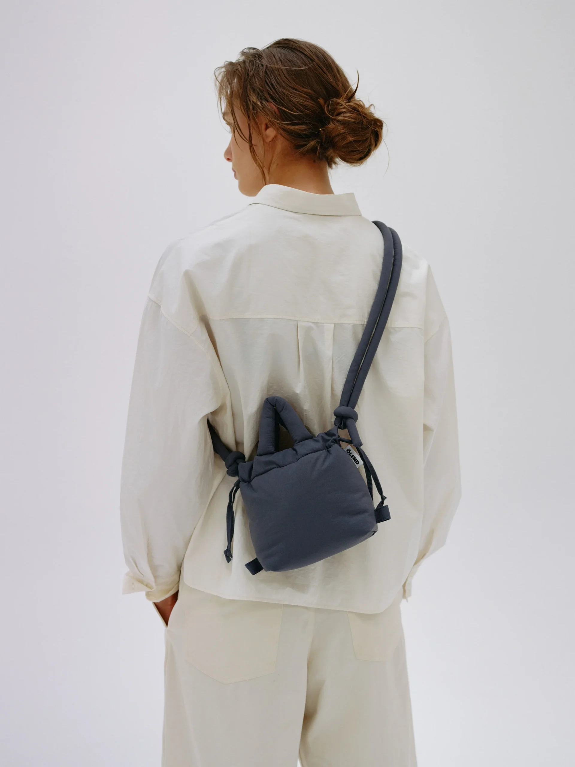 A person with brown hair in a low messy bun, wearing a loose off-white shirt and trousers, stands facing away from the camera. They carry the Micro Ona Soft Bag - Coal, perfect for stylish Tallinn shopping or exploring Estonian design. Gate21.ee