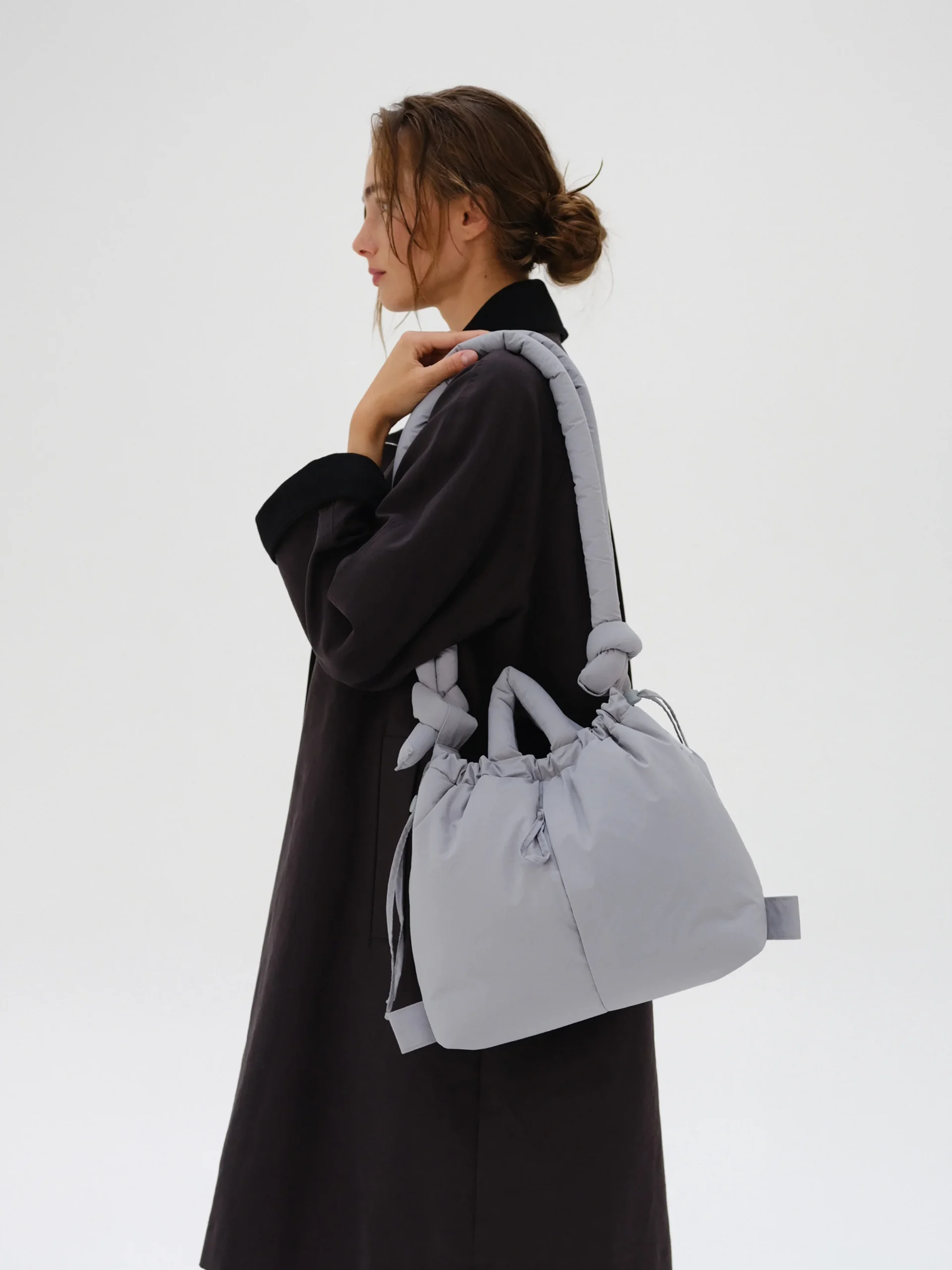 A person with light brown hair in a loose bun, wearing a long dark coat, carries the Ona Soft Bag - Grey—a large, knotted-strap shoulder bag inspired by Estonian design. The profile view is set against a plain white background. Gate21.ee