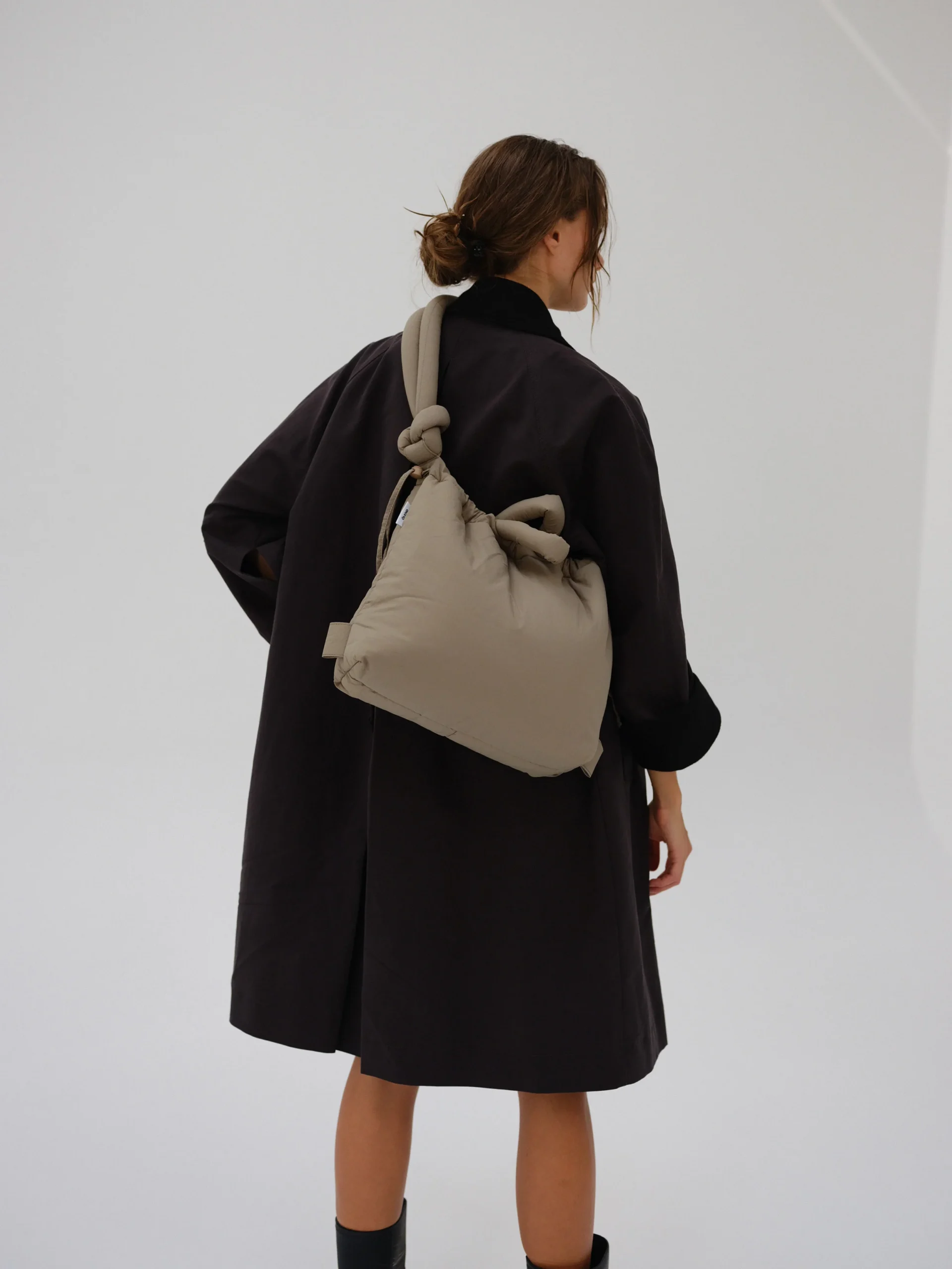 A person with brown hair in a low, messy bun wears a black coat and carries the Ona Soft Bag in Sage—its knotted straps and spacious size make it ideal for Tallinn shopping or picking up finds from an Estonian gift shop—against a plain light background. Gate21.ee