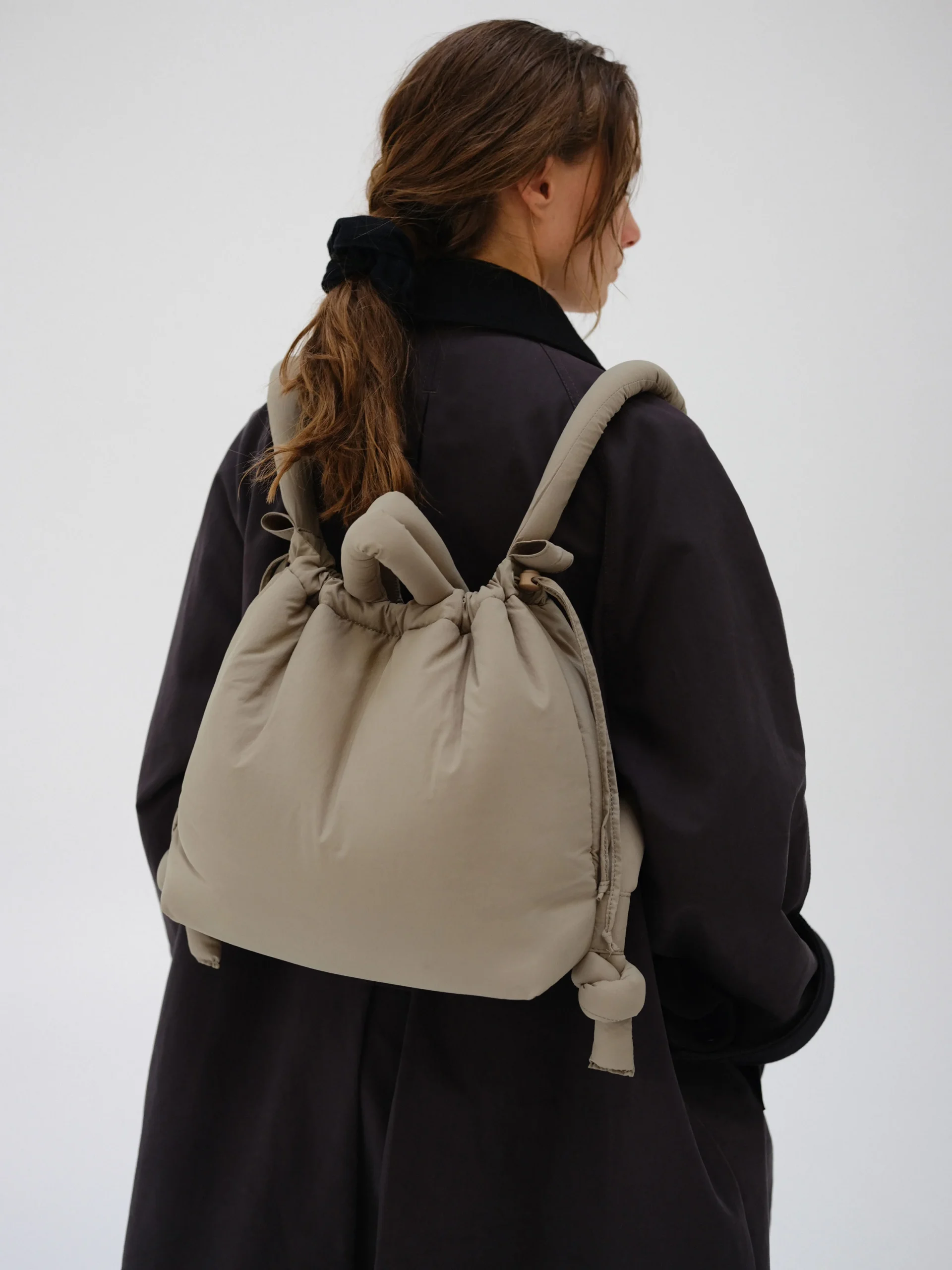 A person with long brown hair in a black tie, wearing a dark coat, carries the Ona Soft Bag - Sage—a large beige fabric rucksack with knot details—ideal for Tallinn shopping or exploring Estonian design. Background is plain and light. Gate21.ee