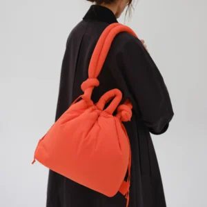 A person with brown hair in a low bun wears a long black coat and carries the Ona Soft Bag - Coral—a large, padded handbag with knotted handles—facing away from the camera against a plain light background. Gate21.ee