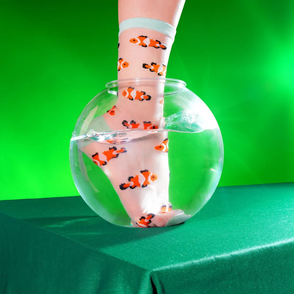 A foot wears Clownfish – Transparent Socks, featuring orange and black fish patterns, submerged in a round fishbowl filled with water on a green surface against a bright green background—showcasing curated Estonian design. Gate21.ee