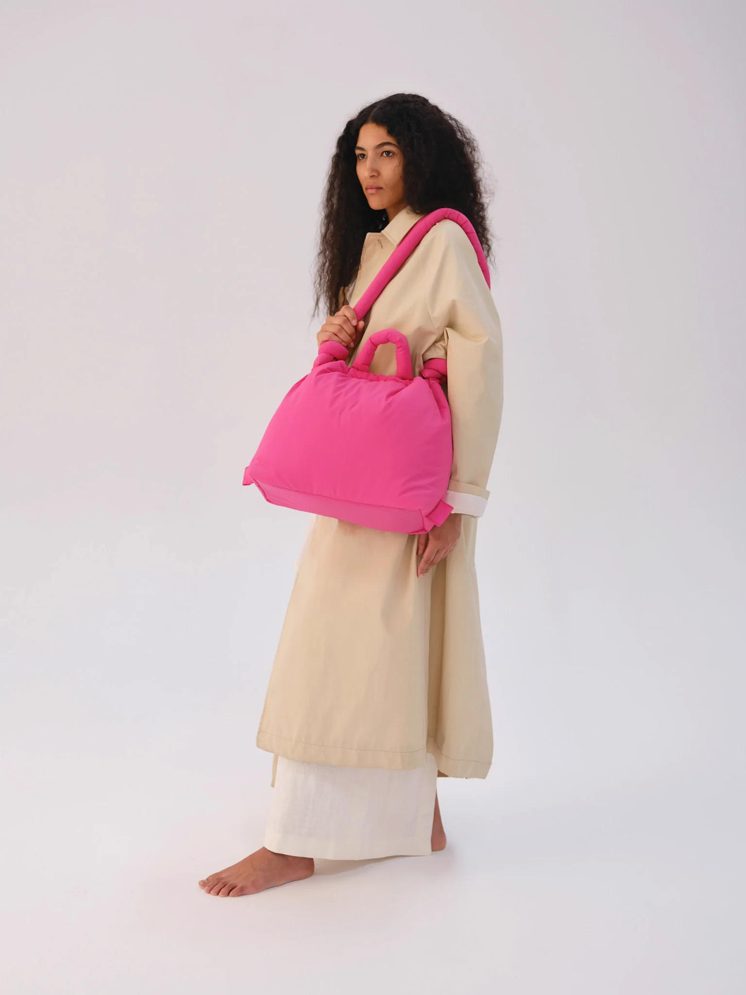 A person with long curly hair stands against a plain background, wearing a long beige outfit and carrying the Ona Soft Bag - Pink—an effortlessly curated look, perfect for shopping in Tallinn or visiting a concept shop. Gate21.ee