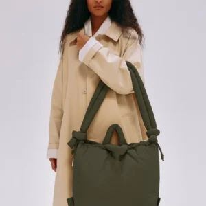 A person in a long beige coat stands against a plain background, holding the Ona Soft Bag - Forest Green with knotted handles—a curated piece of Estonian design ideal for any concept shop. Gate21.ee