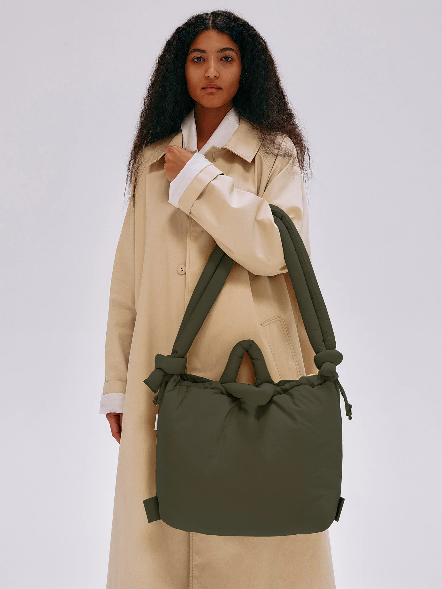 A person in a long beige coat stands against a plain background, holding the Ona Soft Bag - Forest Green with knotted handles—a curated piece of Estonian design ideal for any concept shop. Gate21.ee