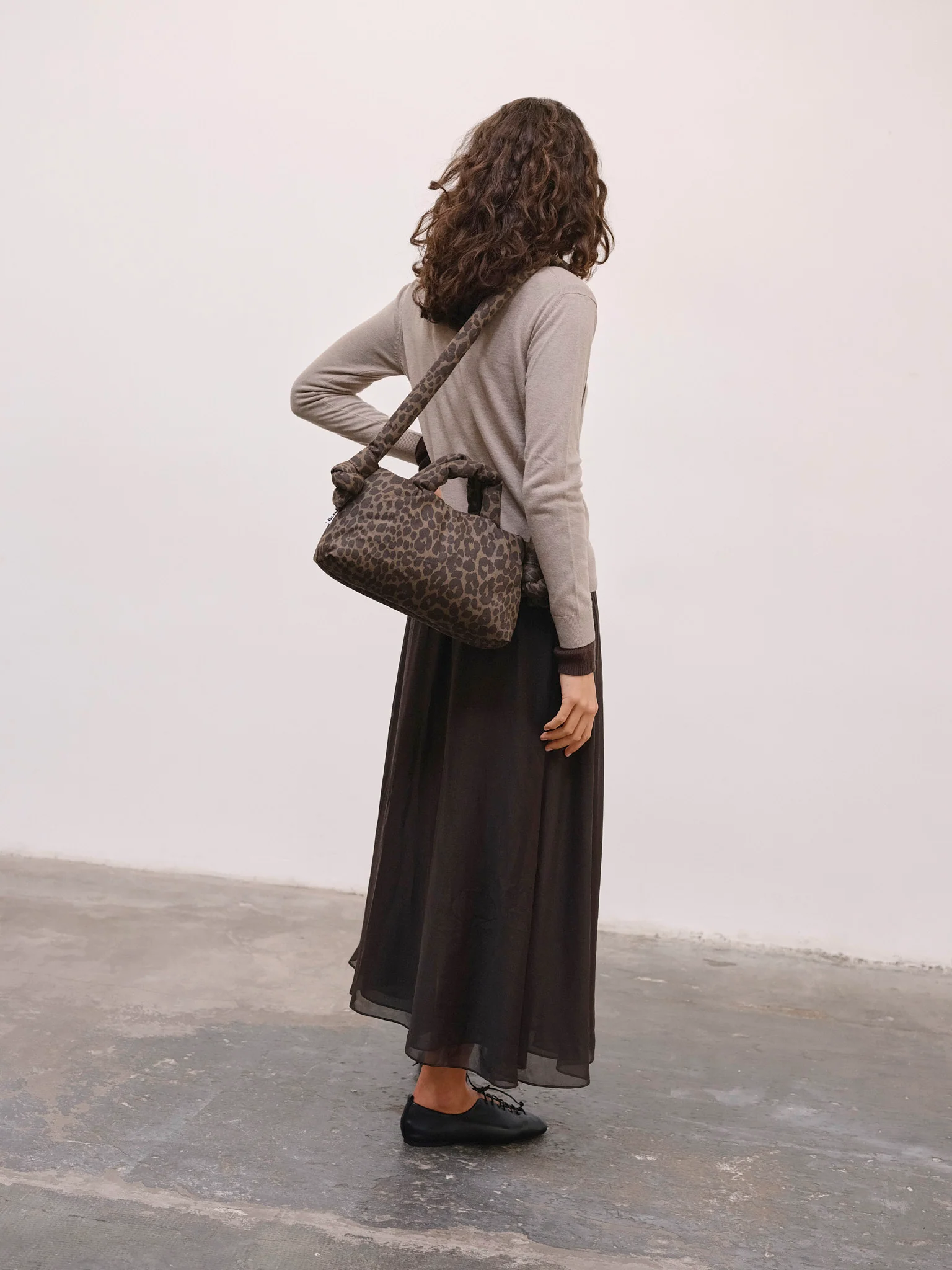 A woman with curly hair faces away, wearing a light long-sleeve top, black maxi skirt, black shoes, and carrying the Mini Ona Soft Bag - Leopard—ideal for curated Tallinn shopping or discovering unique city finds. Gate21.ee