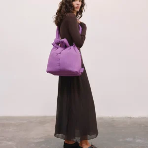 A woman stands indoors against a plain wall, wearing a long black sheer skirt and dark fitted top, carrying the Ona Soft Bag - Grape over one shoulder—a chic look reminiscent of Estonian design gift shops. Gate21.ee