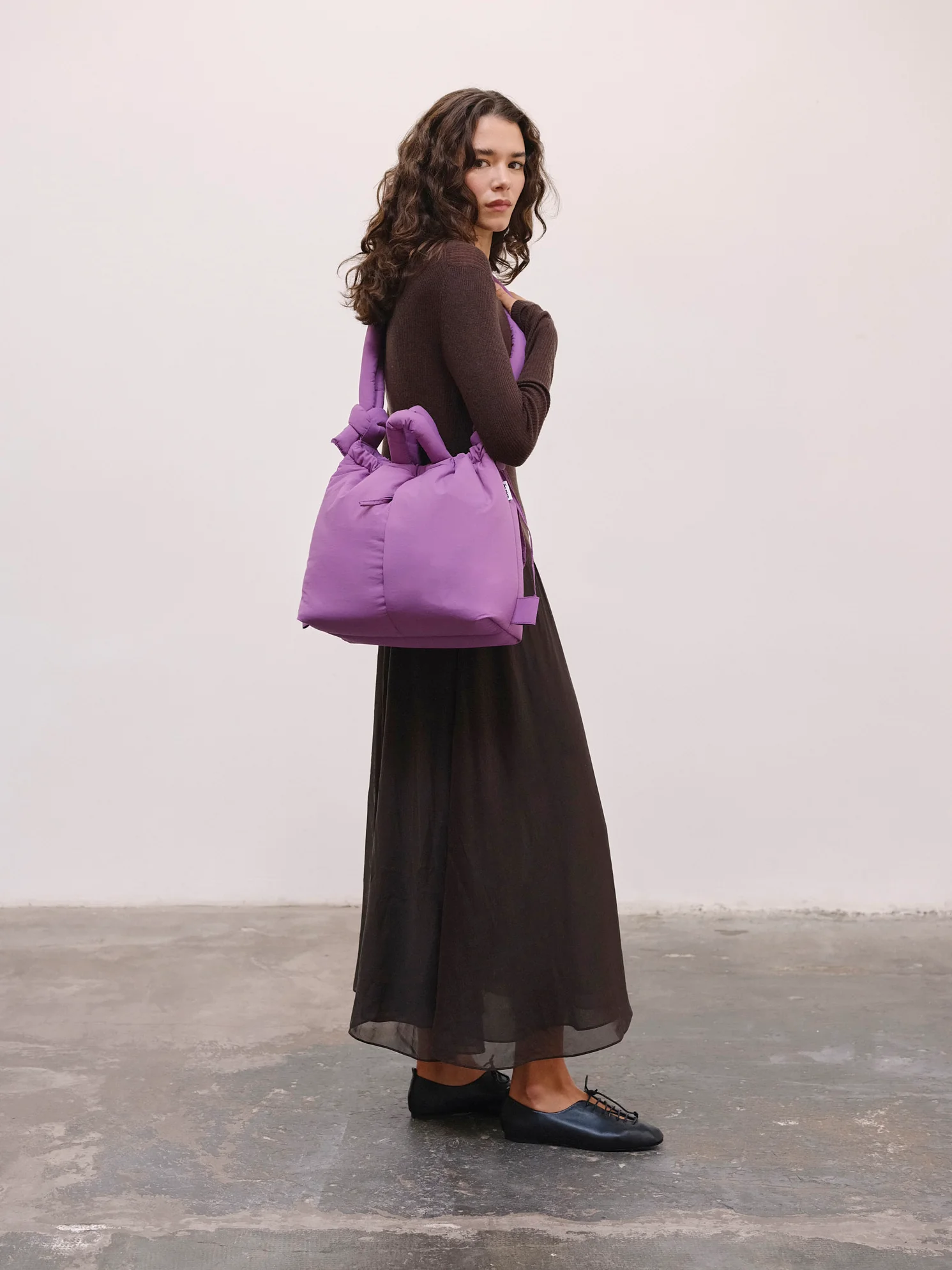 A woman stands indoors against a plain wall, wearing a long black sheer skirt and dark fitted top, carrying the Ona Soft Bag - Grape over one shoulder—a chic look reminiscent of Estonian design gift shops. Gate21.ee
