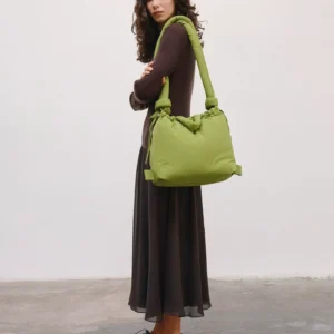 A woman with long curly hair stands on a concrete floor against a plain white wall, wearing a long dark dress, black shoes, and carrying the Ona Soft Bag - Matcha—ideal for discovering curated finds during your Tallinn shopping adventures. Gate21.ee