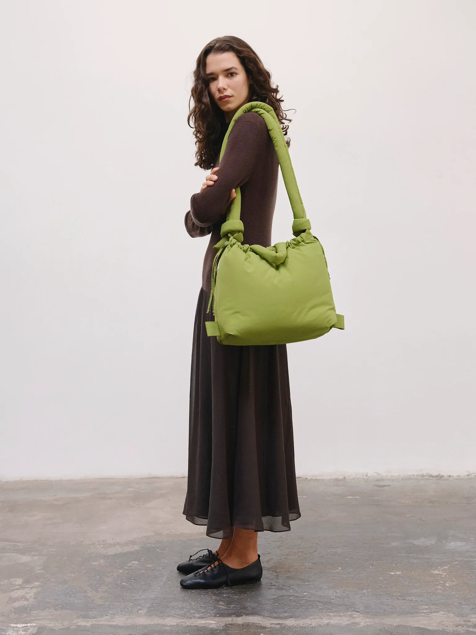 A woman with long curly hair stands on a concrete floor against a plain white wall, wearing a long dark dress, black shoes, and carrying the Ona Soft Bag - Matcha—ideal for discovering curated finds during your Tallinn shopping adventures. Gate21.ee