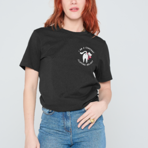 A person with shoulder-length red hair wears a black Fluffing Delight T-Shirt, featuring white text and a small graphic. They stand against a plain light background, gazing to the side with one hand on their hip. Gate21.ee