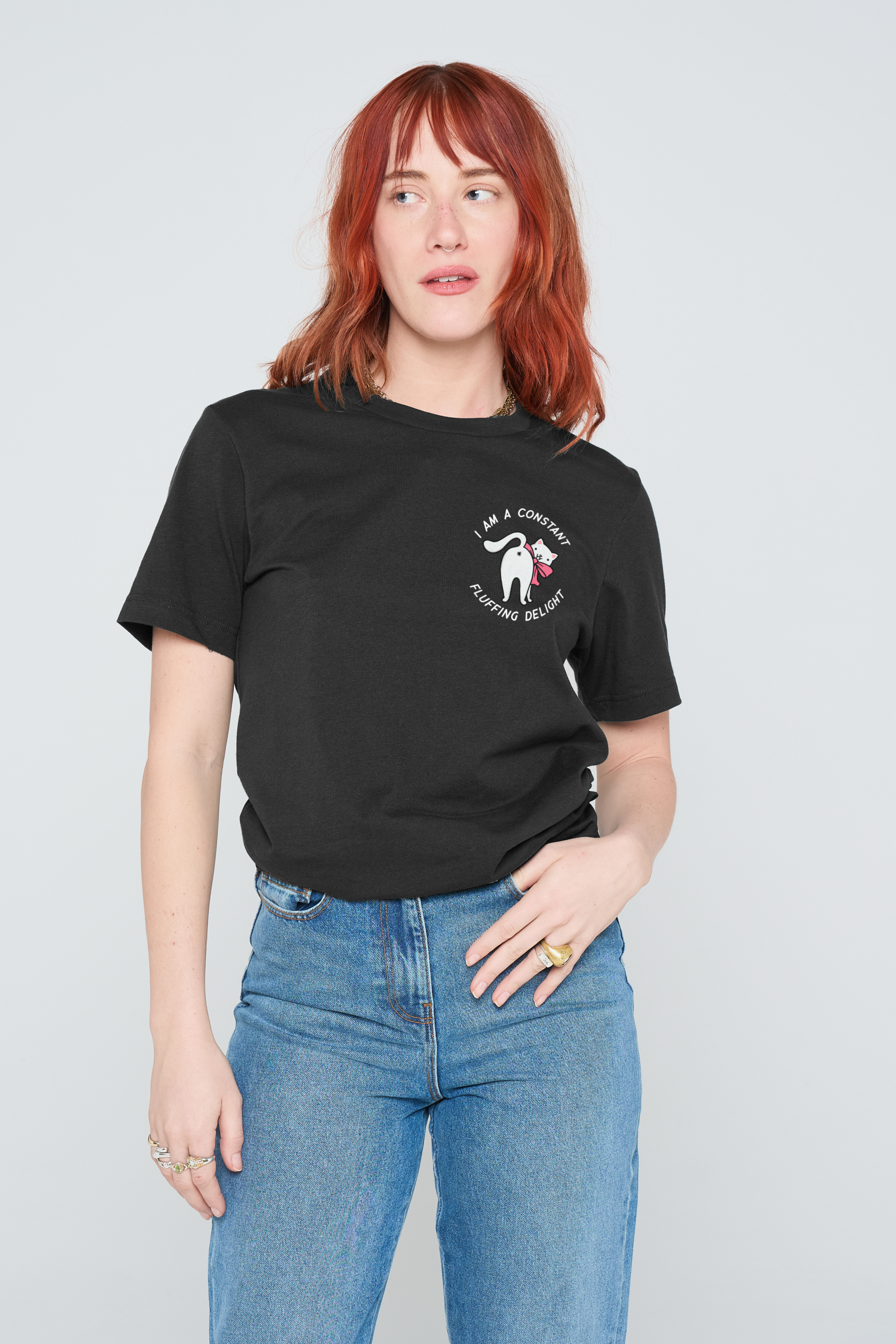 A person with shoulder-length red hair wears a black Fluffing Delight T-Shirt, featuring white text and a small graphic. They stand against a plain light background, gazing to the side with one hand on their hip. Gate21.ee