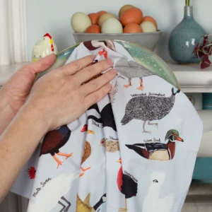 A person dries a plate with the Fowl - Tea Towel, featuring charming bird motifs. Nearby, a bowl of eggs and a ceramic chicken sit beside a blue vase—a stylish home scene perfect for any curated gift shop. Gate21.ee