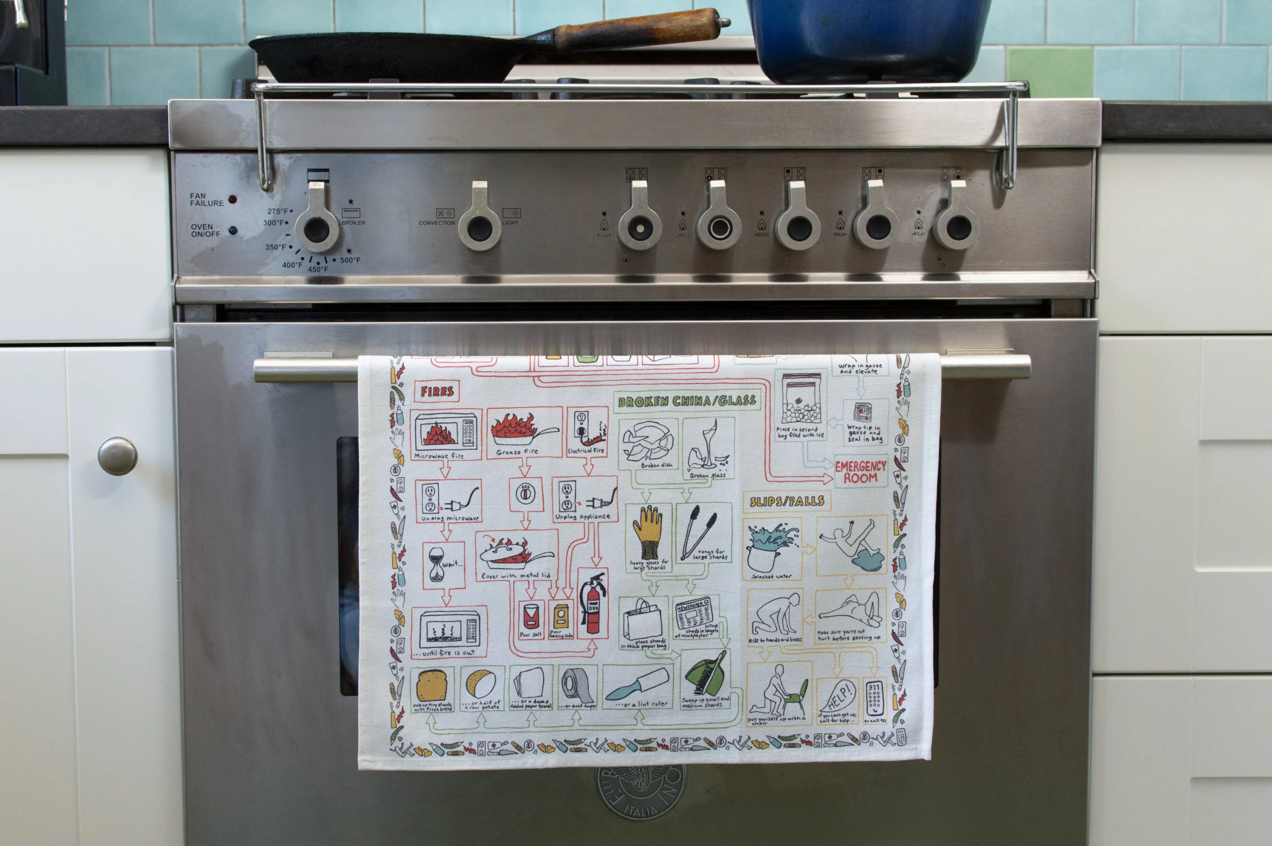 A stainless steel oven with five front control knobs displays the Kitchen Accidents – Tea Towel, known for its colourful, curated illustrations. The kitchen features a light blue tile splashback and white cupboards—ideal for Tallinn shopping inspiration. Gate21.ee