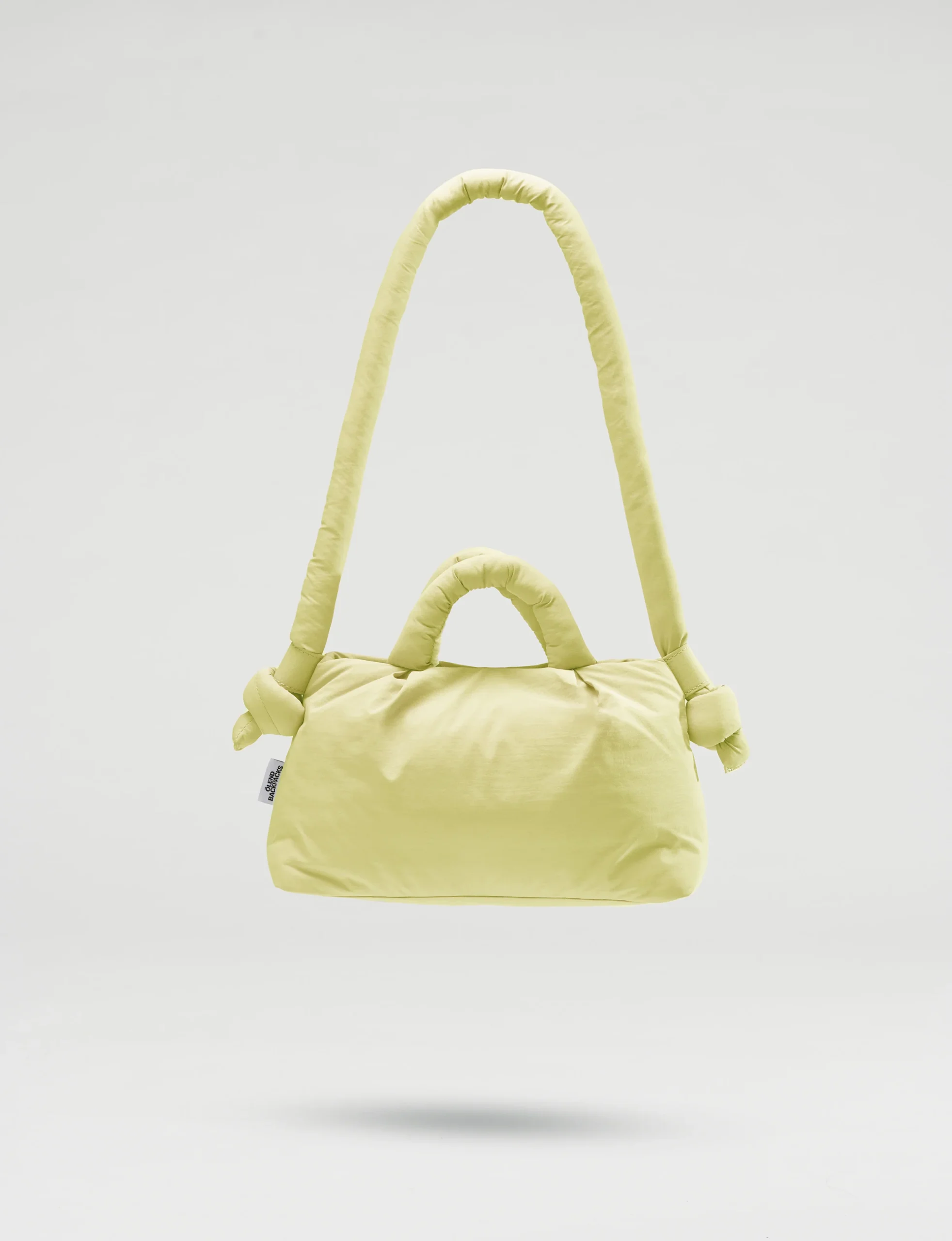 The Mini Ona Soft Bag - Lime is a light yellow padded handbag with short handles and a long, knotted shoulder strap, displayed against a plain light grey background—a chic, plush accessory for stylish shoppers. Gate21.ee