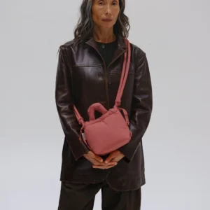 A person with long grey hair wears a dark brown leather jacket and dark trousers, carrying the Micro Ona Soft Bag in Soft Peach—an understated nod to Estonian design. The background is plain and light-coloured. Gate21.ee
