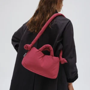 A person with wet, wavy hair wears a long black coat and carries the Mini Ona Soft Bag in Cherry Red, featuring knotted handles and a thick strap, standing against a plain light background. Gate21.ee