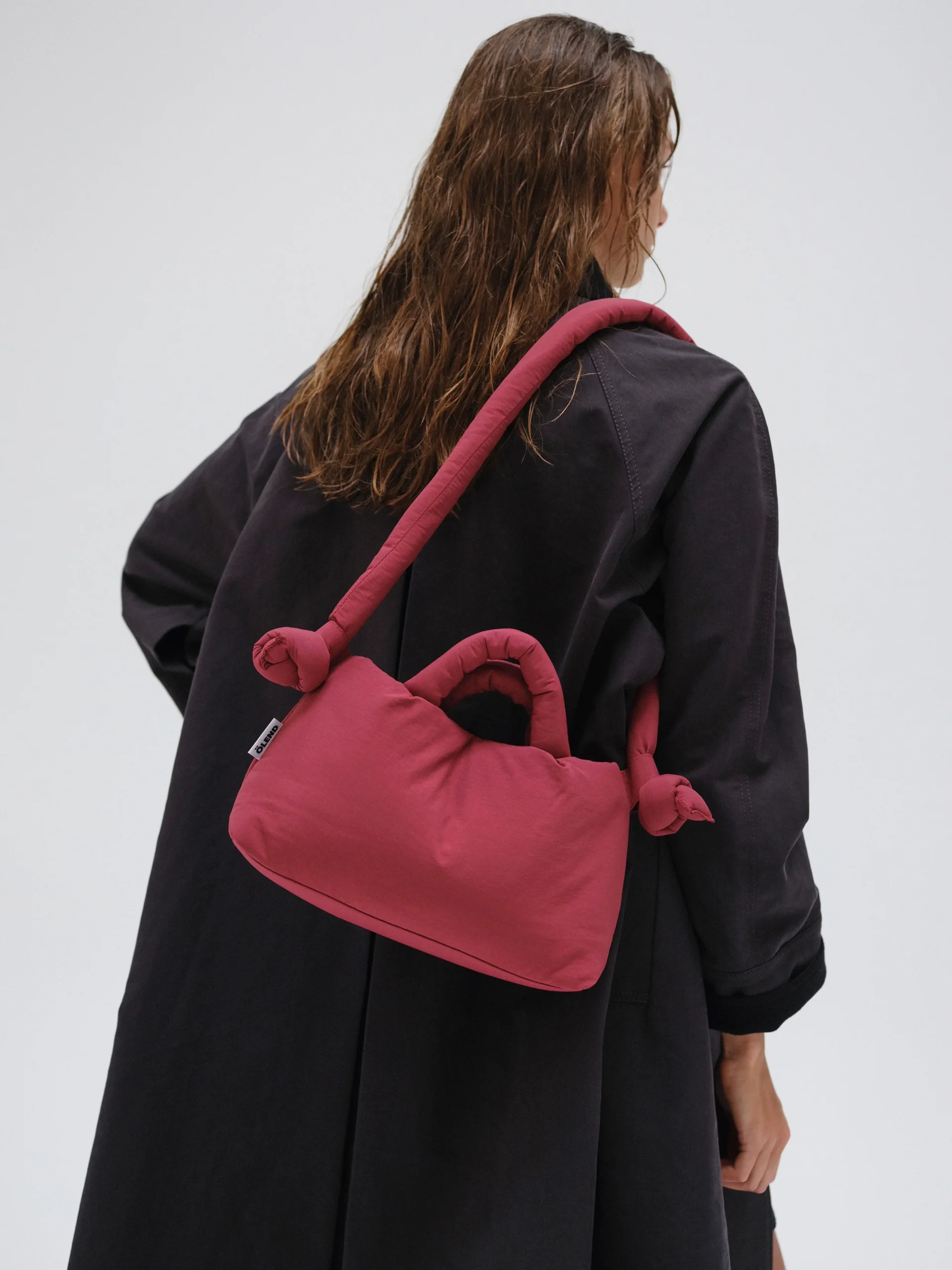 A person with wet, wavy hair wears a long black coat and carries the Mini Ona Soft Bag in Cherry Red, featuring knotted handles and a thick strap, standing against a plain light background. Gate21.ee