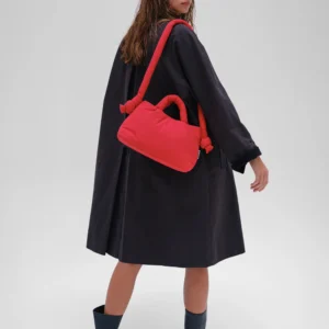 A woman in a loose, dark coat and boots stands angled away, glancing over her shoulder with the Mini Ona Soft Bag - Red slung across her body—a stylish nod to Estonian design. The background is plain and light-coloured. Gate21.ee