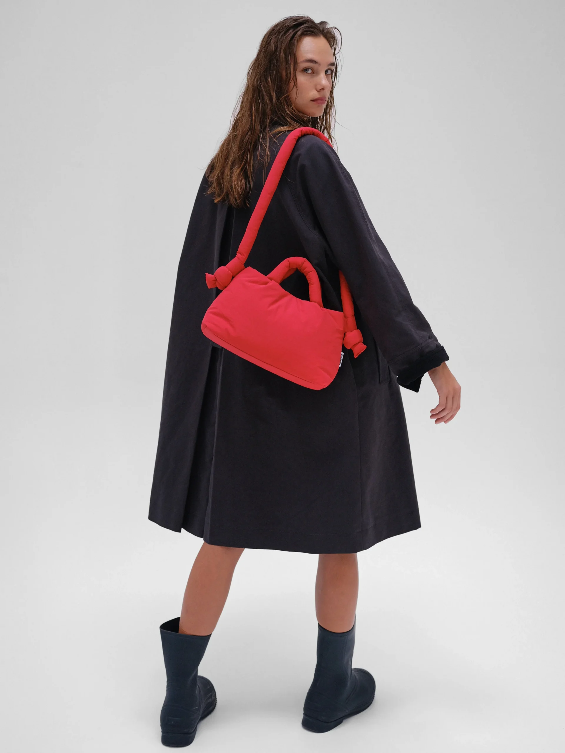 A woman in a loose, dark coat and boots stands angled away, glancing over her shoulder with the Mini Ona Soft Bag - Red slung across her body—a stylish nod to Estonian design. The background is plain and light-coloured. Gate21.ee