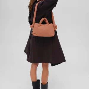 A person faces away, wearing a long dark coat, black boots, and a black hair accessory, carrying the Mini Ona Soft Bag in Soft Peach—a unique design inspired by Estonia. The background is plain and light grey. Gate21.ee