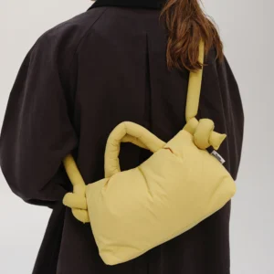 A person with long brown hair tied back wears a dark oversized coat and carries the Mini Ona Soft Bag in Lime—an eye-catching, padded handbag with knotted straps inspired by Estonian design. The photo is taken from behind, showing their back and side profile. Gate21.ee