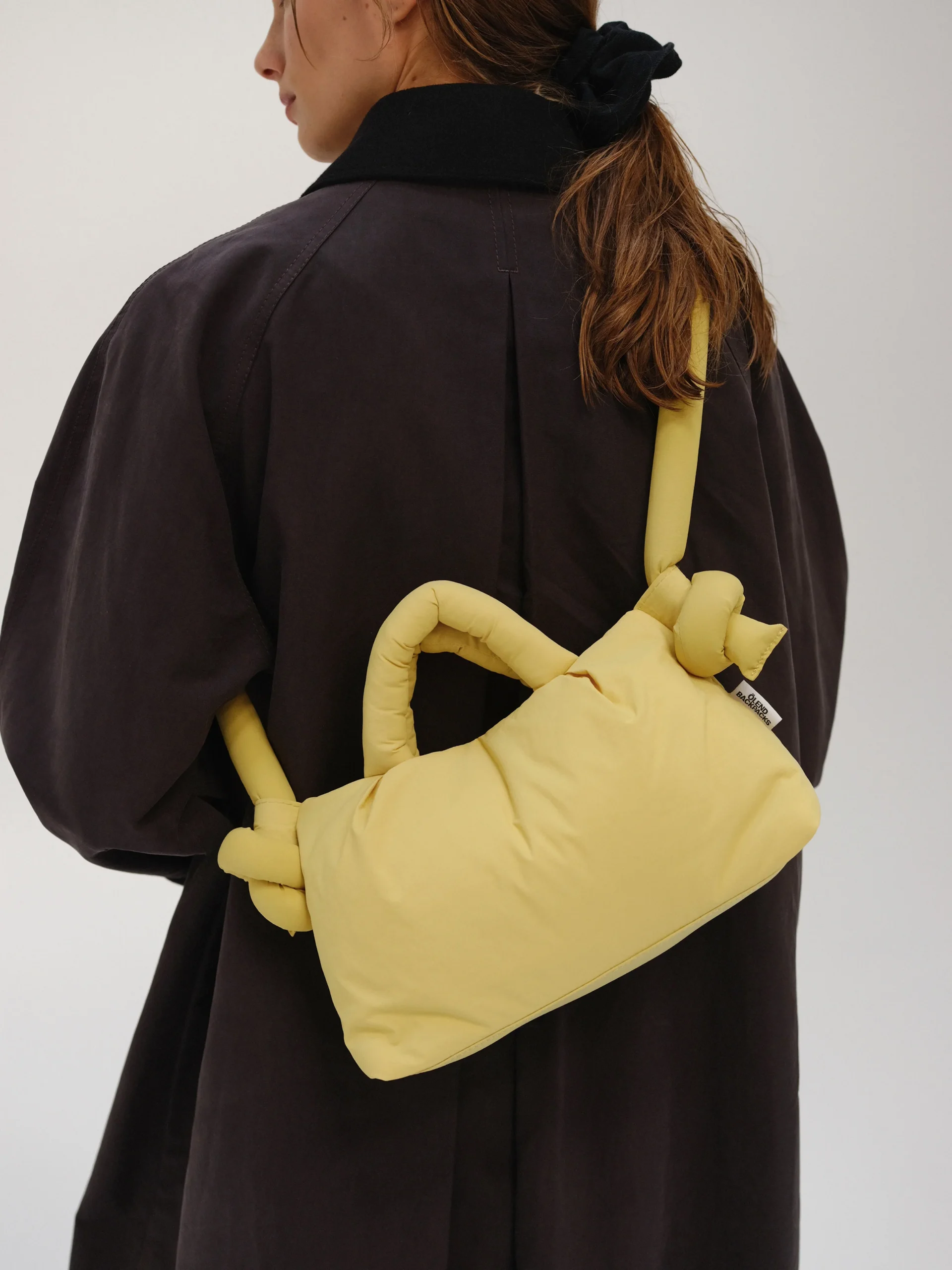 A person with long brown hair tied back wears a dark oversized coat and carries the Mini Ona Soft Bag in Lime—an eye-catching, padded handbag with knotted straps inspired by Estonian design. The photo is taken from behind, showing their back and side profile. Gate21.ee