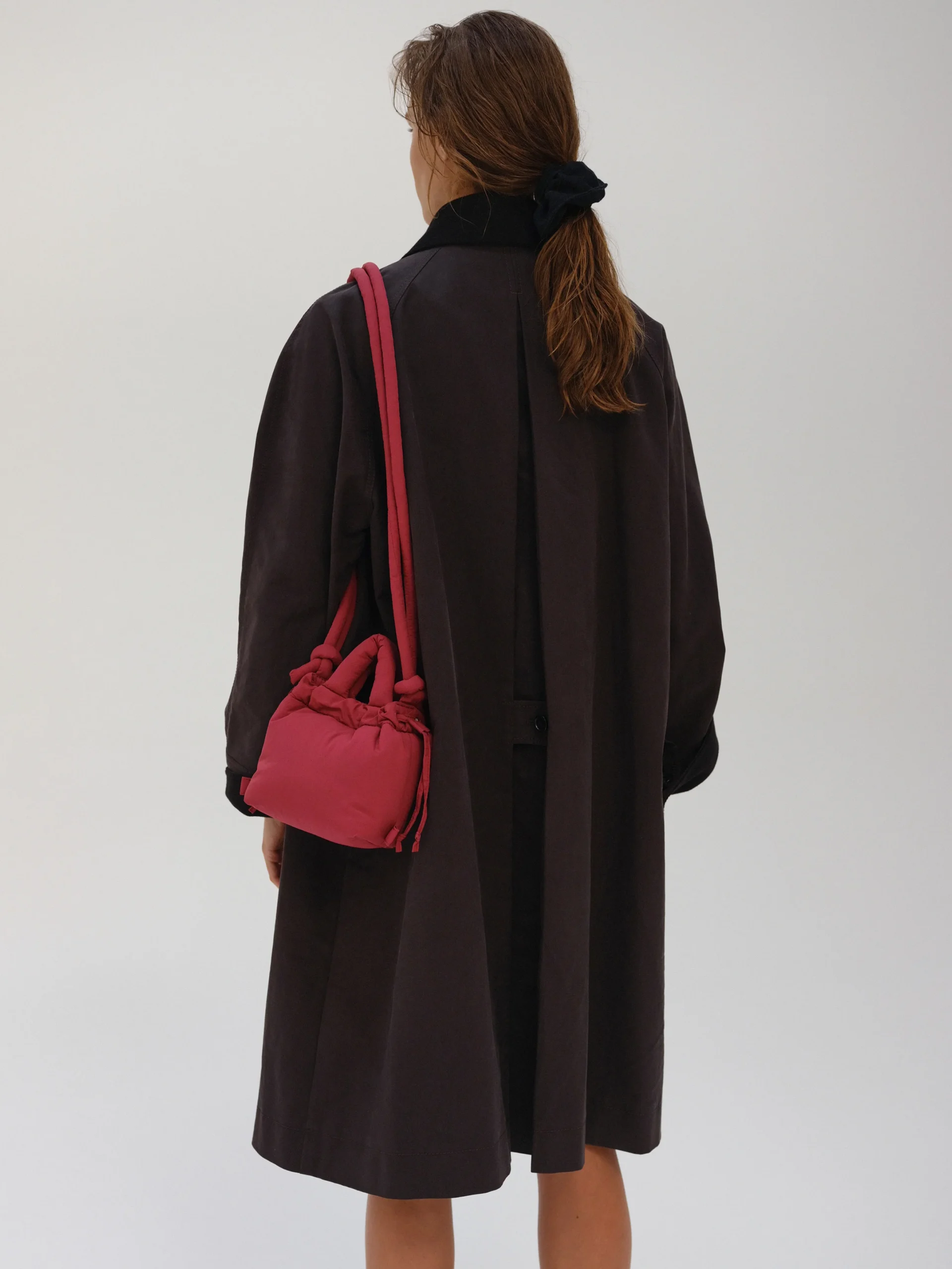A person with long brown hair tied back, dressed in a dark coat, carries the Micro Ona Soft Bag in Cherry Red over their shoulder. They face away from the camera against a plain background, highlighting this standout gift shop find. Gate21.ee