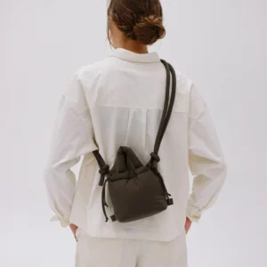 Seen from behind, a person with brown hair in a low bun wears an off-white shirt and trousers, carrying the Micro Ona Soft Bag in Forest Green. The small drawstring pouch adds a touch of curated style against a plain, light background. Gate21.ee