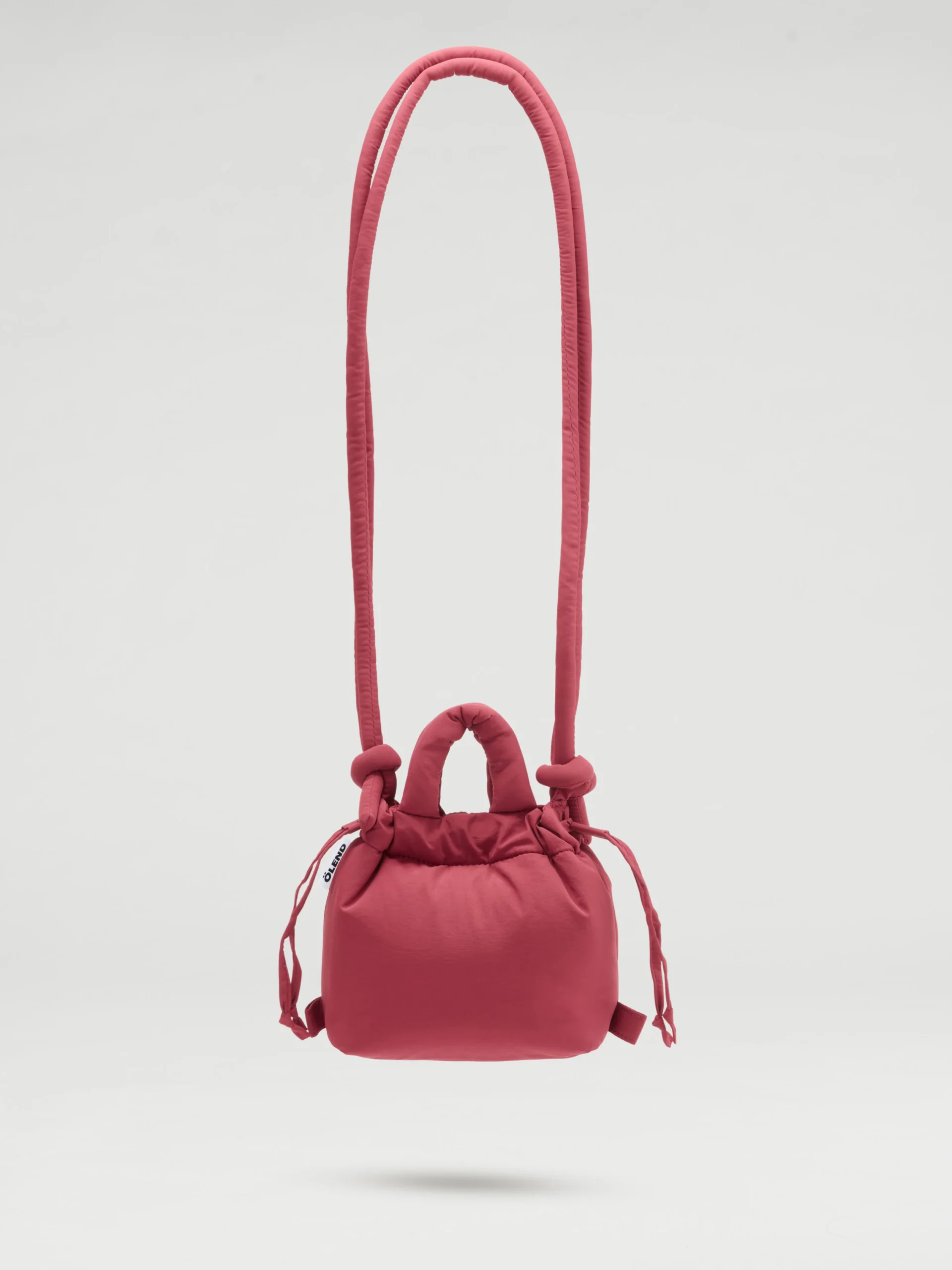The Micro Ona Soft Bag - Cherry Red, a small square-shaped handbag with a plush finish, padded handle, long shoulder strap, and side drawstrings, showcases curated Estonian design against a plain white background. Gate21.ee