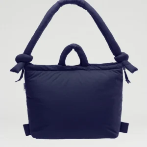 The Ona Soft Bag - Navy features a padded, rectangular design with a wide handle, two knotted details on each end, and a smaller centre handle. Its modern look makes it a standout choice for gift shops against any light background. Gate21.ee
