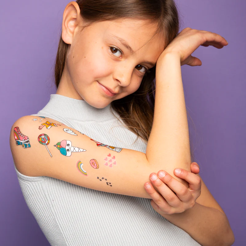 A young girl wearing a sleeveless light grey top poses against a purple background, showing off colourful Lily - Temporary Tattoos on her shoulder and upper arm at a trendy Tallinn concept shop. Gate21.ee