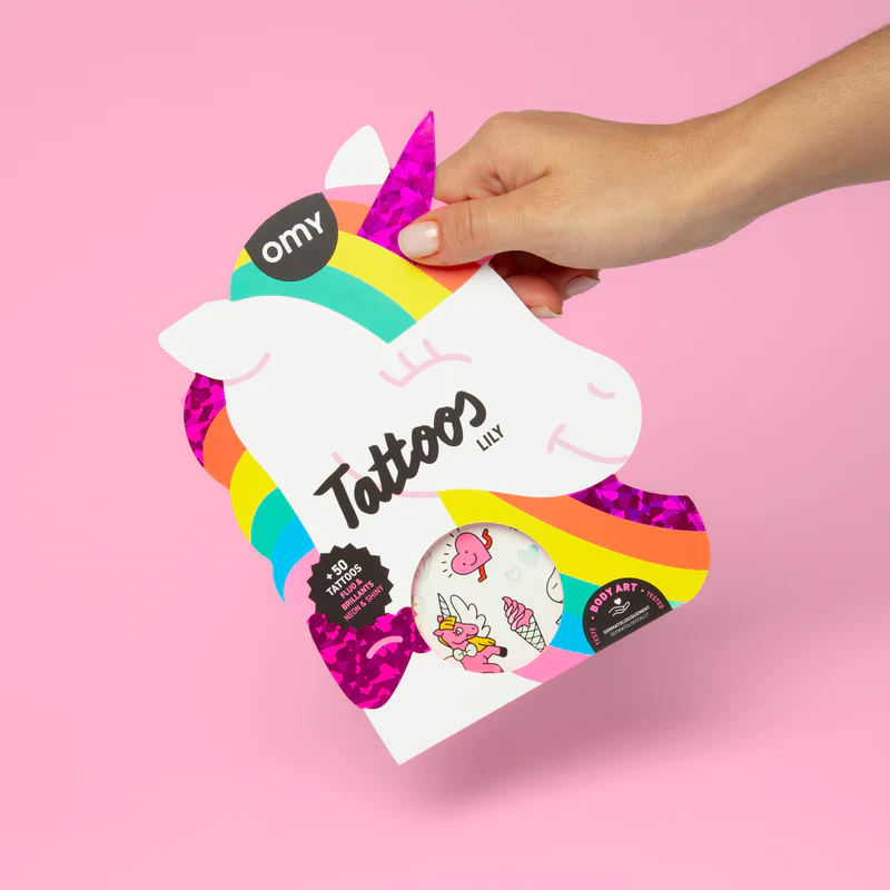 A hand holds the Lily - Temporary Tattoos unicorn-shaped packaging with colourful rainbow accents and "Tattoos" text against a pink background, offering a peek at playful designs—ideal for any gift shop display. Gate21.ee