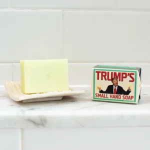 A bar of Trump - Soap rests on a soap dish beside its box, which features an illustrated man in a suit, all placed on a white tiled bathroom surface. Gate21.ee