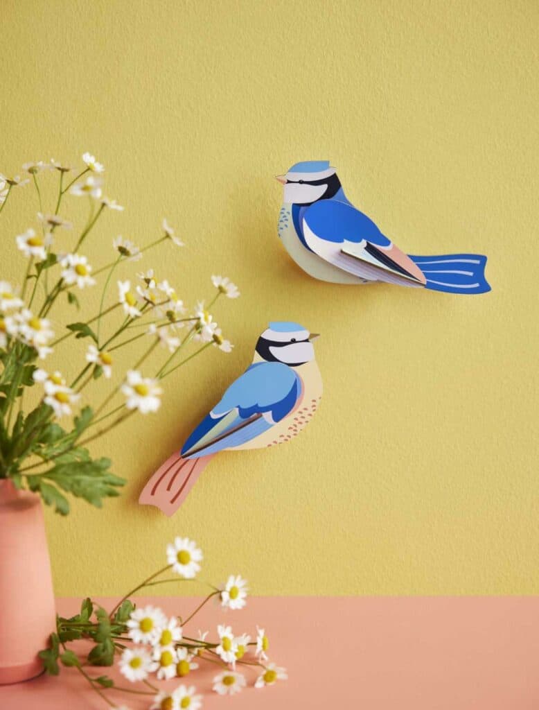 A curated vase with white daisies sits on a pink surface before a yellow wall. Above, two Blue Tits—colourful paper birds—add charm and unique inspiration for any gift shop display. Gate21.ee