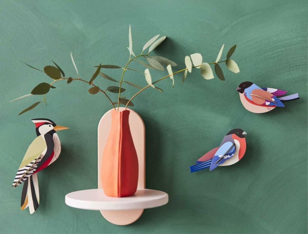 A decorative wall shelf displays a vase with foliage against a green background. Next to it, three colourful "Woodpecker" paper bird sculptures add Estonian design charm, evoking the vibe of a Tallinn concept shop. Gate21.ee