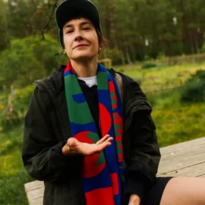A person in a dark jacket, baseball cap, and colourful scarf sits outdoors on a wooden surface amid greenery, tossing an organic tomato—perhaps after exploring curated Tallinn shopping or visiting a local gift shop. Gate21.ee